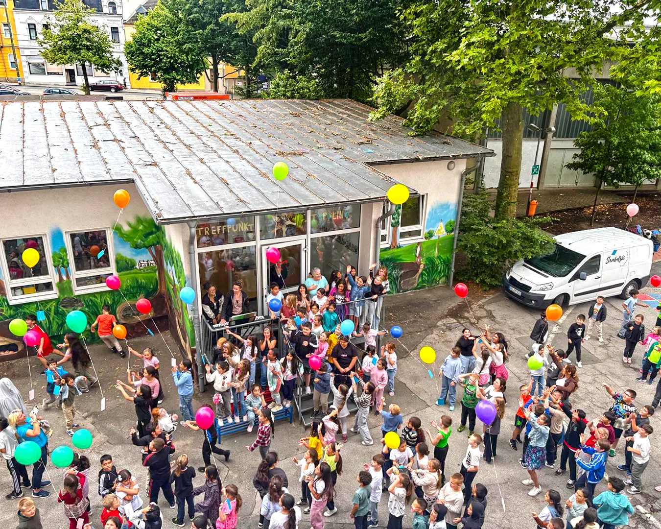 Auf dem Bild sieht man viele Kinder mit Luftballons. Sie stehen auf dem Schulhof. An den Luftballons hängen Karten mit Wünschen für den Frieden.